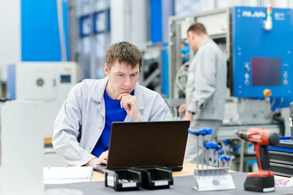 Worker reviews data on a laptop at a desk in an industrial workspace while another person works in the background.