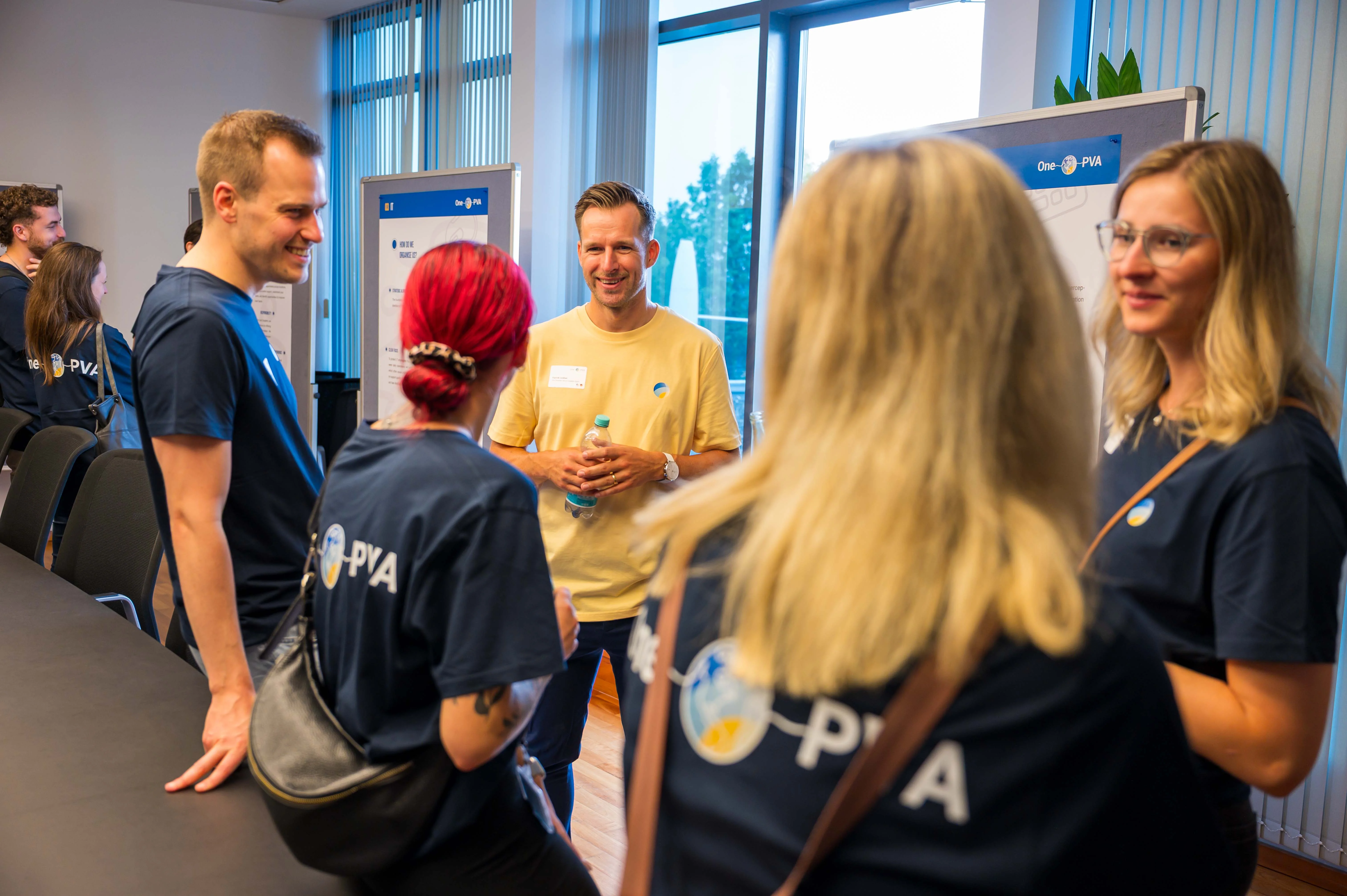 Group of PVA employees in a modern office or meeting room, engaged in conversation. Several people wear dark blue PVA shirts, with presentation boards and large windows in the background