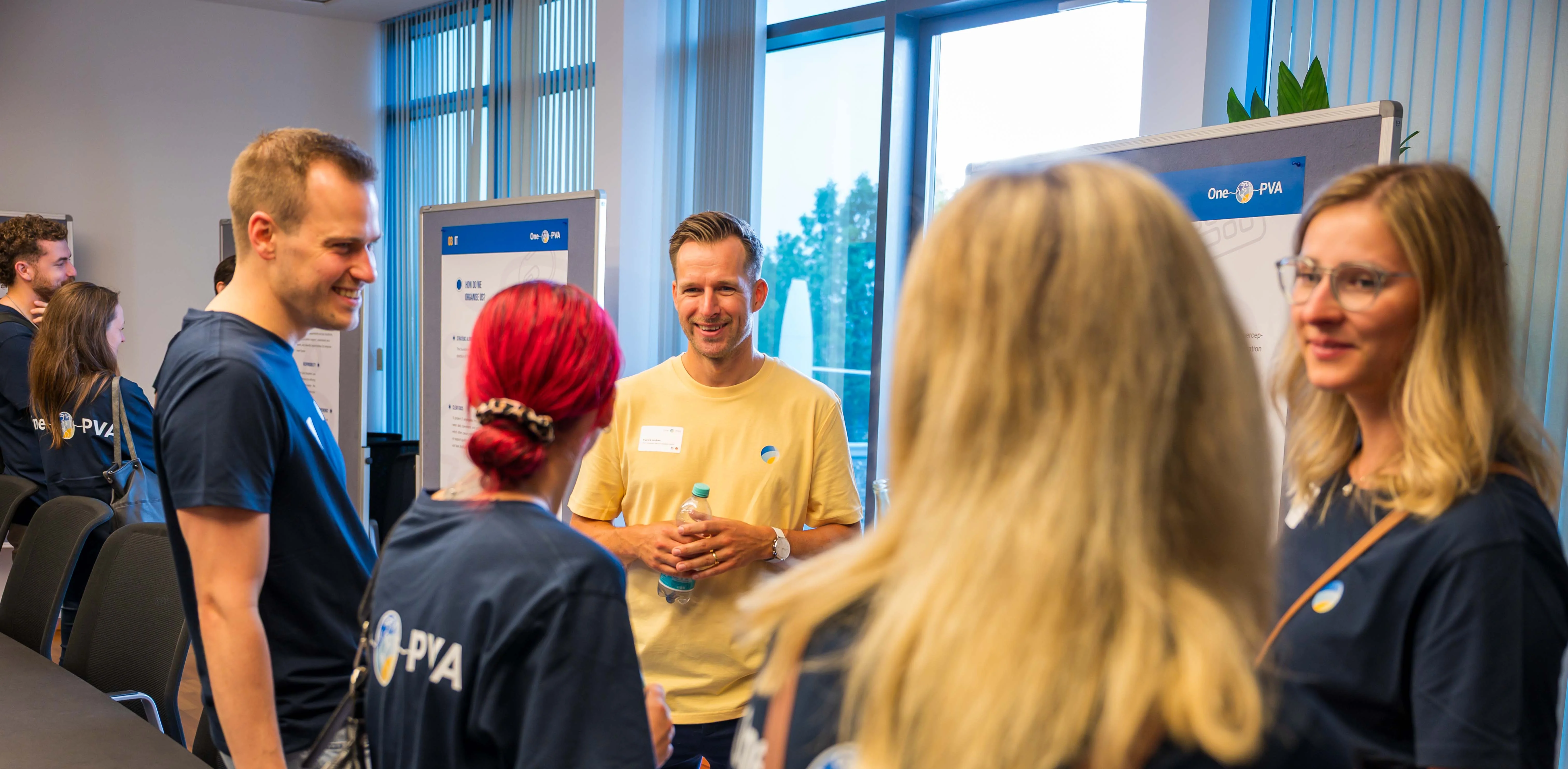 Group of PVA employees in a modern office or meeting room, engaged in conversation. Several people wear dark blue PVA shirts, with presentation boards and large windows in the background