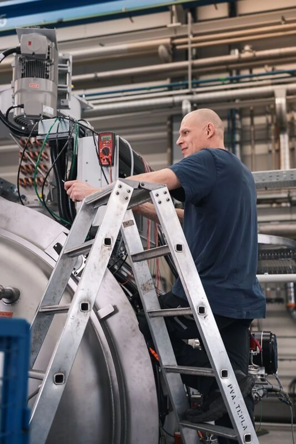 Worker on a ladder adjusts components on a large industrial machine inside a spacious production hall.