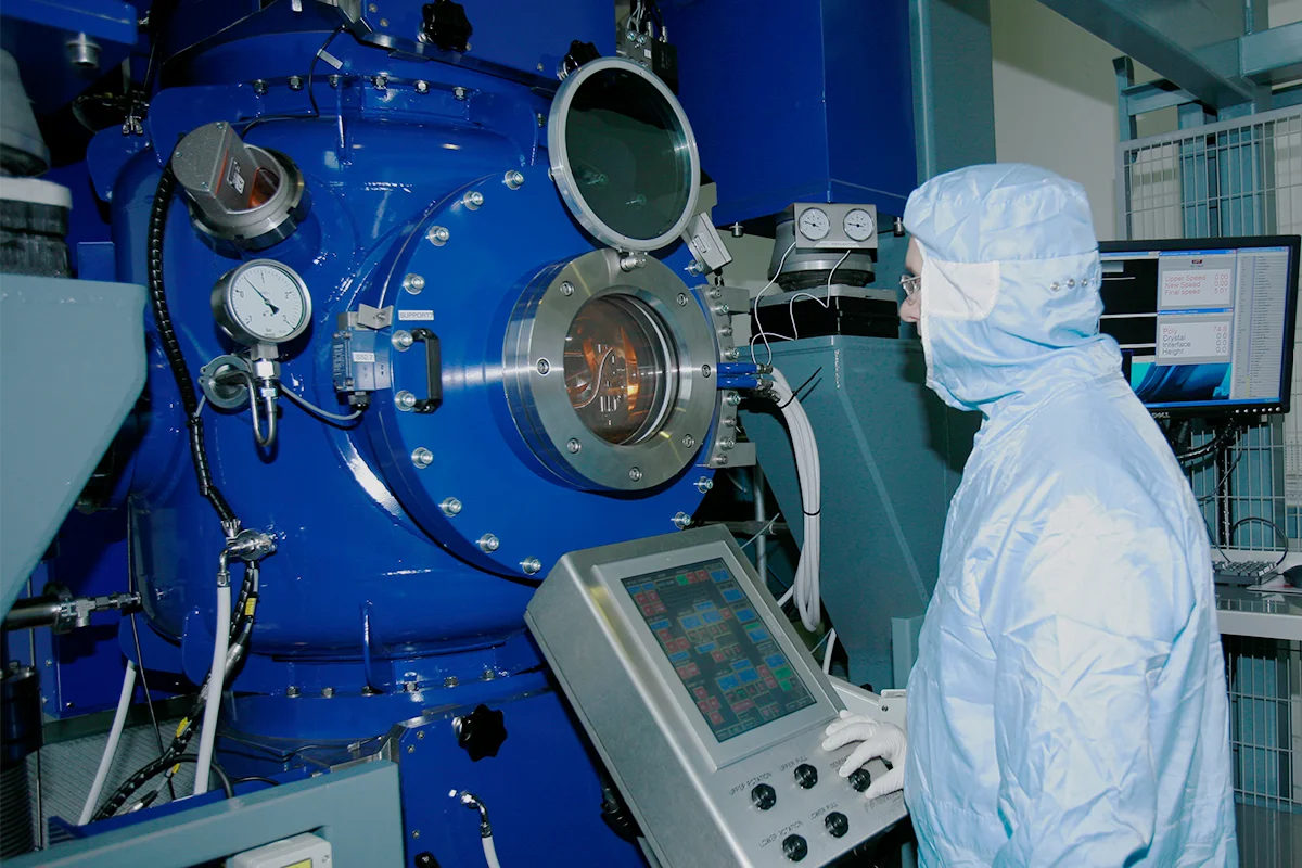 Operator in cleanroom suit monitoring a float‑zone crystal growth system through the observation window