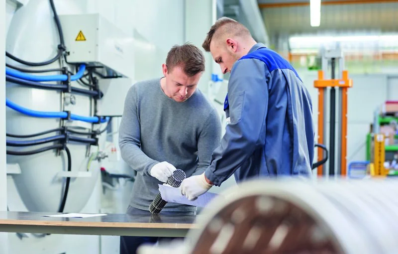 Two workers examine metal parts on a table, discussing details in a bright industrial workshop.