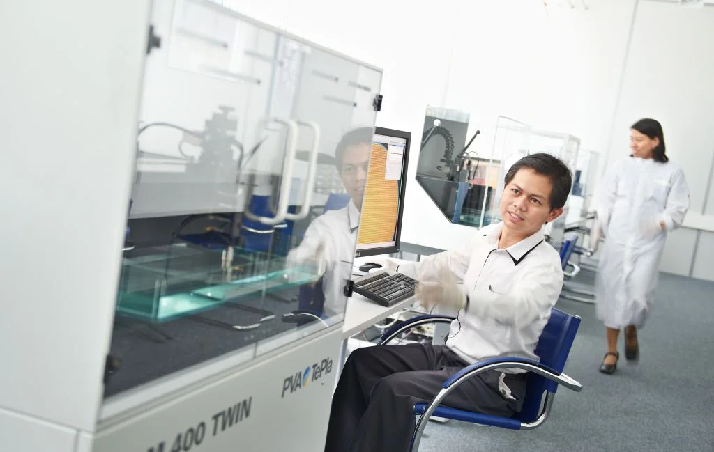 Lab worker operates scanning acoustic microscopy process in a clean, bright laboratory while another person walks in the background.