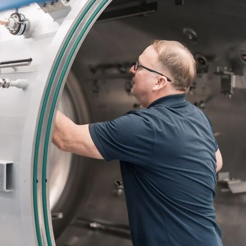 Worker inspects the interior of a large vacuum chamber, checking components and fittings.