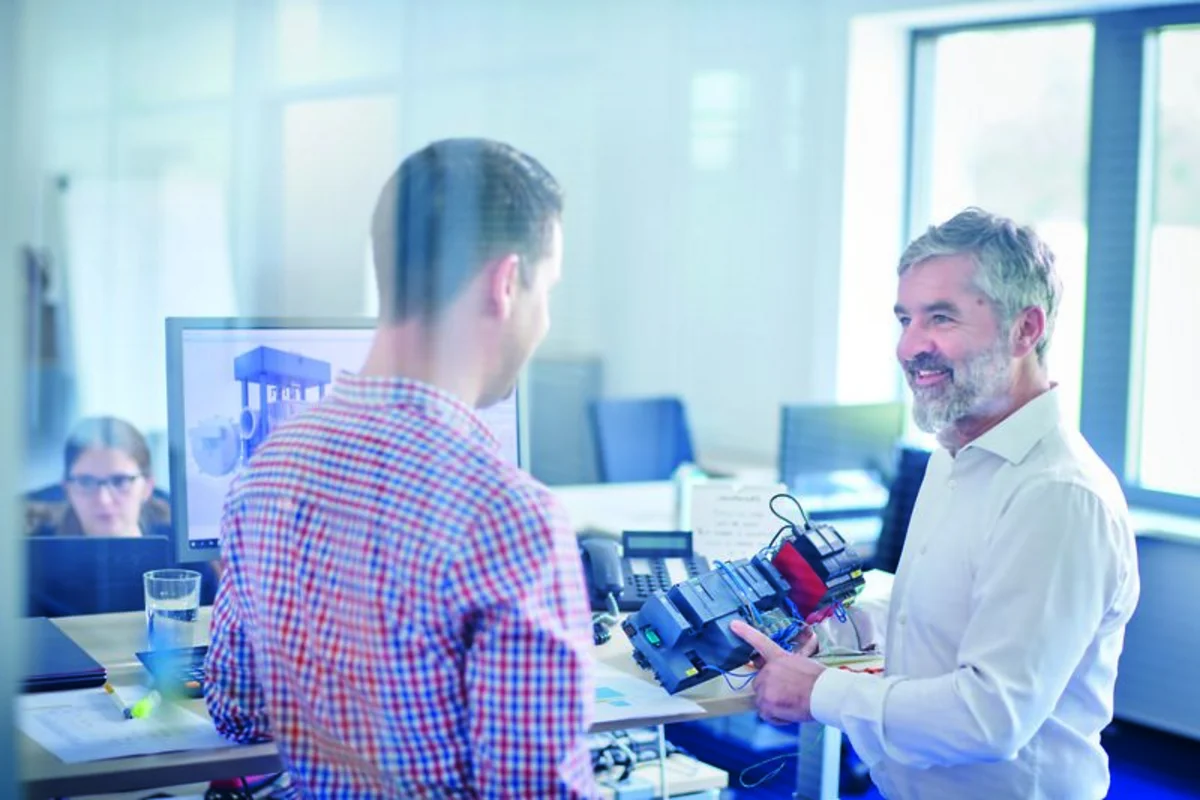 Two colleagues discuss technical equipment in a bright office while another person works at a computer nearby.