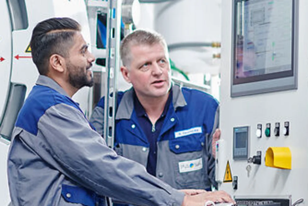 Two workers operate a control panel beside a large industrial furnace, discussing machine settings.